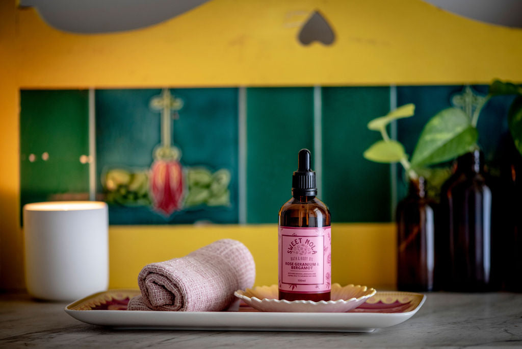 Rose geranium and bergamot bath and body oil. In 100ml glass amber bottle with pink label. It sits on a ceramic dish with a cotton face washer.